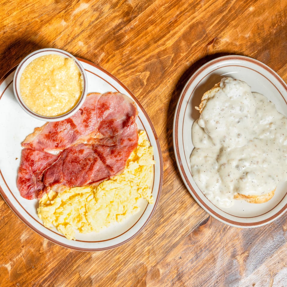 Scrambled eggs, ham, and grits on one plate, with biscuits and gravy on another, on a wooden table.