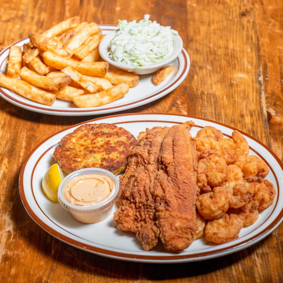 Fried fish, shrimp, and crab cake with fries and coleslaw on a rustic wooden table.